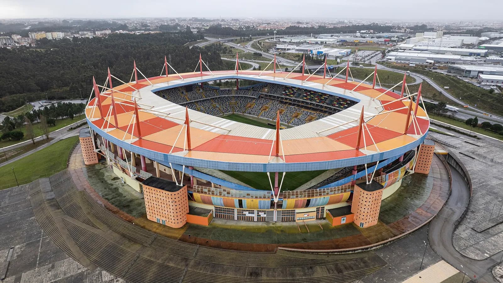 Aerial view of an oval stadium with a roof in bands of red, orange and pink, held up by red masts and cables, and perforated orange towers at the corners, set among pine woods, roads and warehouses.