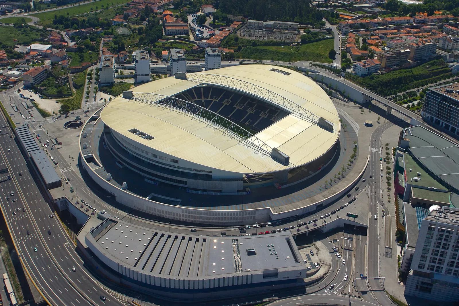 Aerial view of a large oval stadium with a pale roof that leaves the centre open over the pitch, two long steel arches running across the roof, surrounded by motorways, car parks, a low flat-roofed building and apartment blocks.