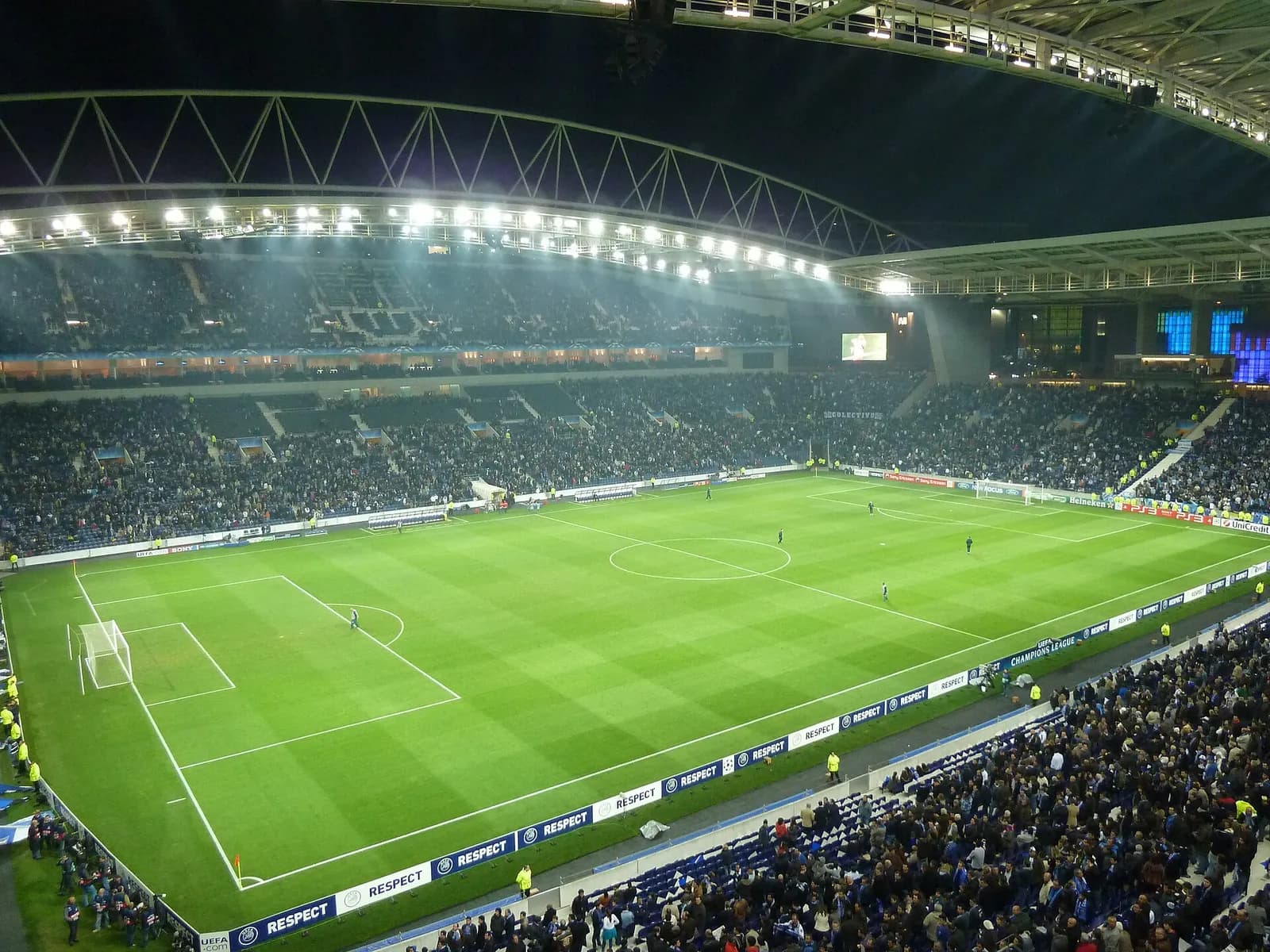 Floodlit interior of a football stadium at night: a green pitch with a few figures on it, two tiers of stands full of spectators, and a white steel truss along the edge of the roof.