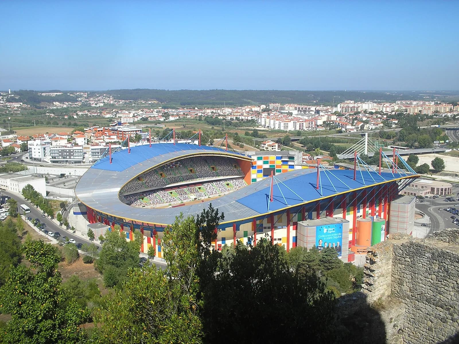 View from a stone castle wall over trees to a stadium with a blue roof on red masts and a façade of multicoloured squares, with apartment blocks and a cable-stayed road bridge behind.