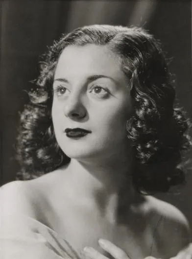Black and white studio portrait of a young woman with dark curled hair to her shoulders and dark lipstick, looking up and to the left, one hand at her collarbone.