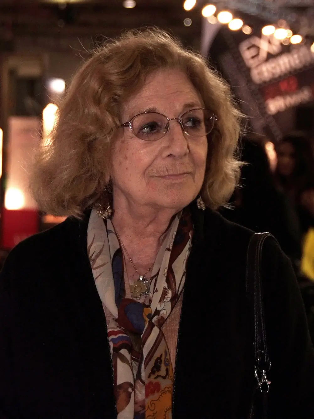 An older woman with wavy shoulder-length fair hair and glasses, wearing a dark coat and a patterned scarf, looks off to one side with a slight smile. Out-of-focus lights glow behind her.