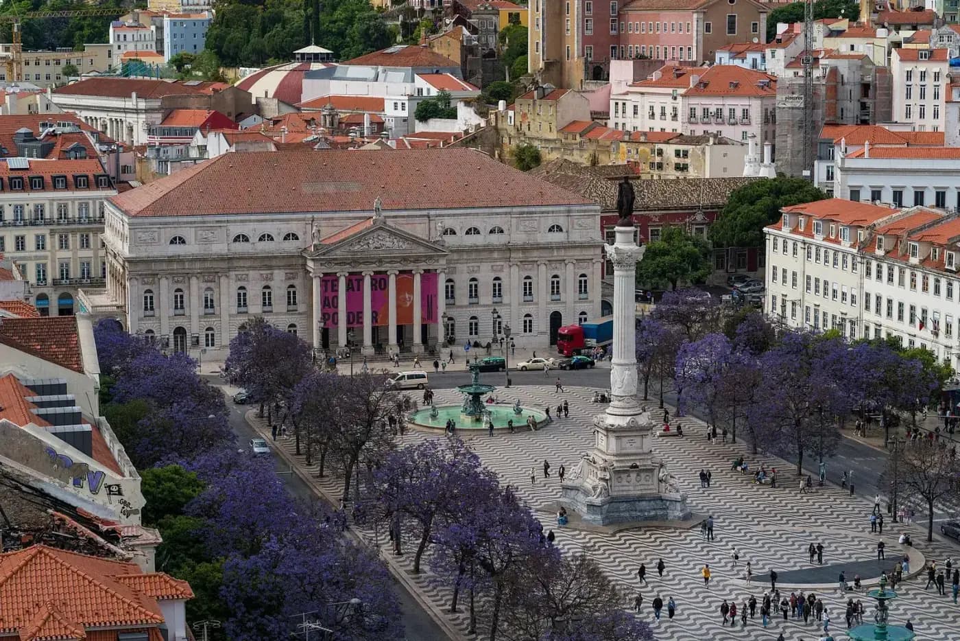 A view from above of Lisbon's Rossio square, paved in wavy black and white stone, with jacaranda trees in purple bloom, a tall column topped by a statue, and fountains. At the far end stands a long neoclassical building with a columned portico hung with red banners, the national theatre.