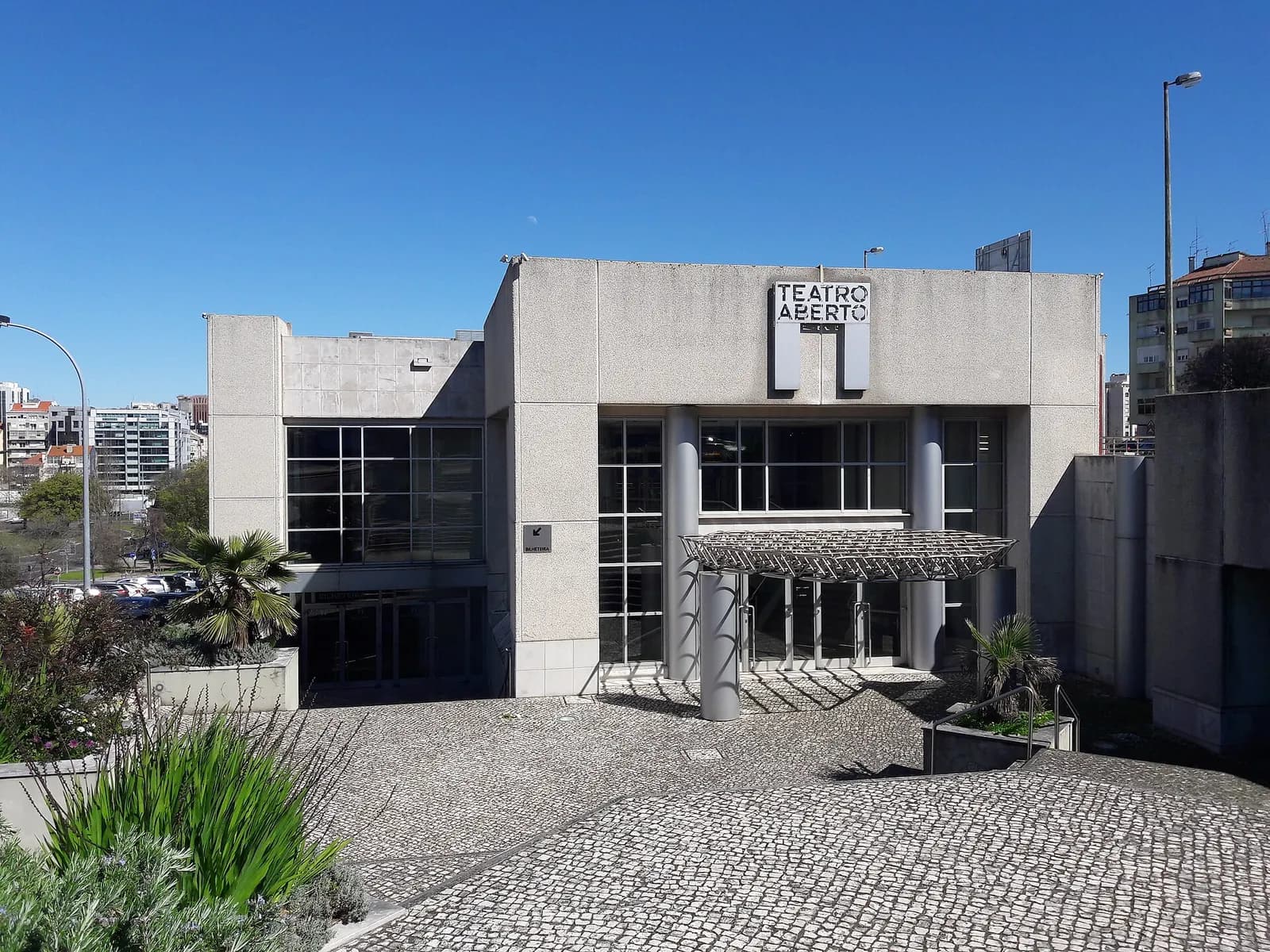 The plain concrete and glass front of a modern theatre building, its name in black letters on a white panel, set back behind a stone-paved forecourt with plants and a palm tree.