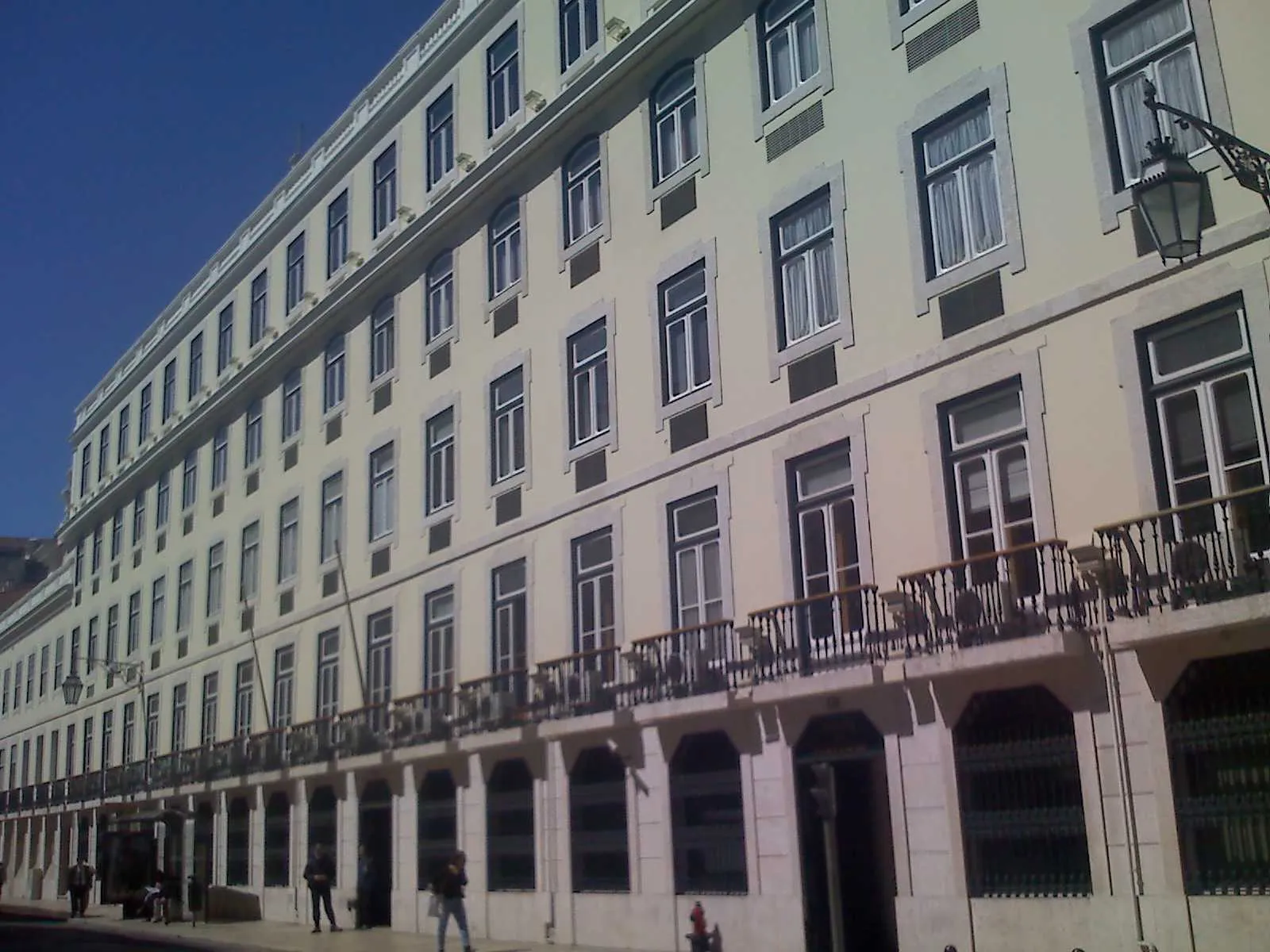 A long pale yellow building with rows of stone-framed windows, a balcony with iron railings along the first floor and arched doorways at street level, seen at an angle under a deep blue sky. A few people walk on the pavement.