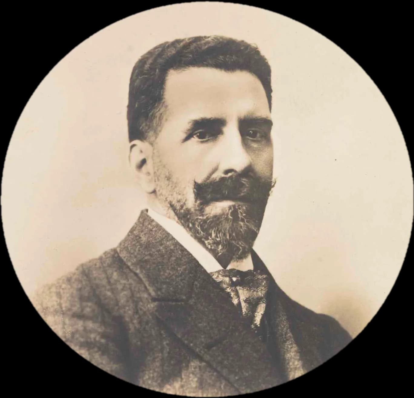 Sepia studio portrait in a round frame of a man with short dark hair, a thick moustache and a greying beard, wearing a tweed jacket and a patterned tie.