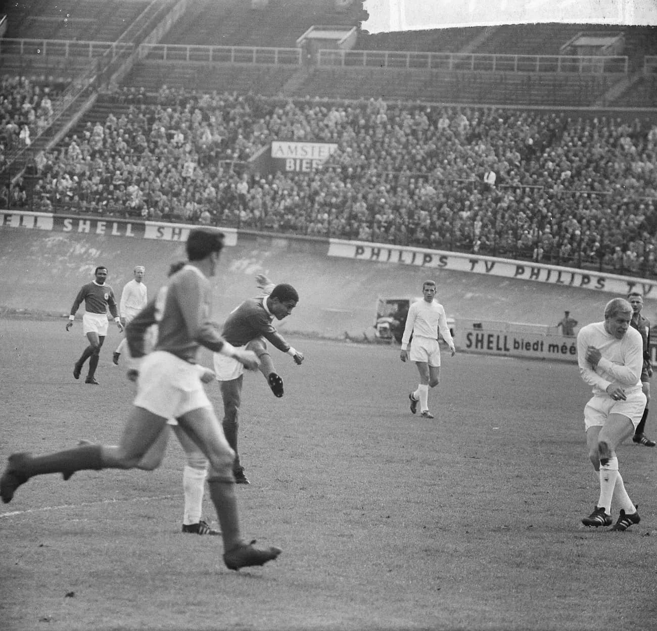 A player in a dark Benfica shirt follows through on a shot in the middle of the pitch while a defender in white on the right turns away, arms folded across his chest. Other players, advertising hoardings and a large crowd in the stands fill the background.