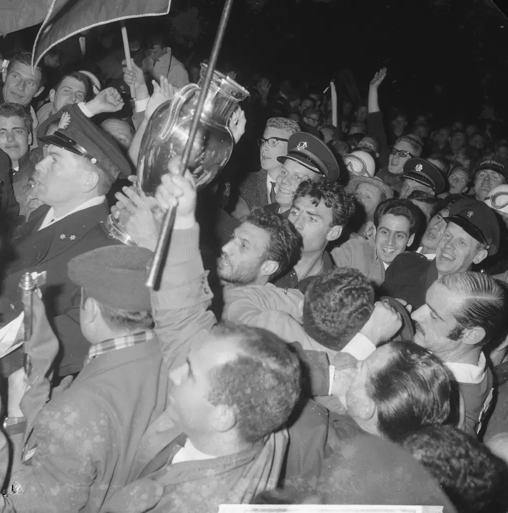 A dense, jubilant crowd at night presses around a silver trophy with large handles held up above their heads. Uniformed policemen in peaked caps are caught in the crush, and a flag on a pole leans over the scene.