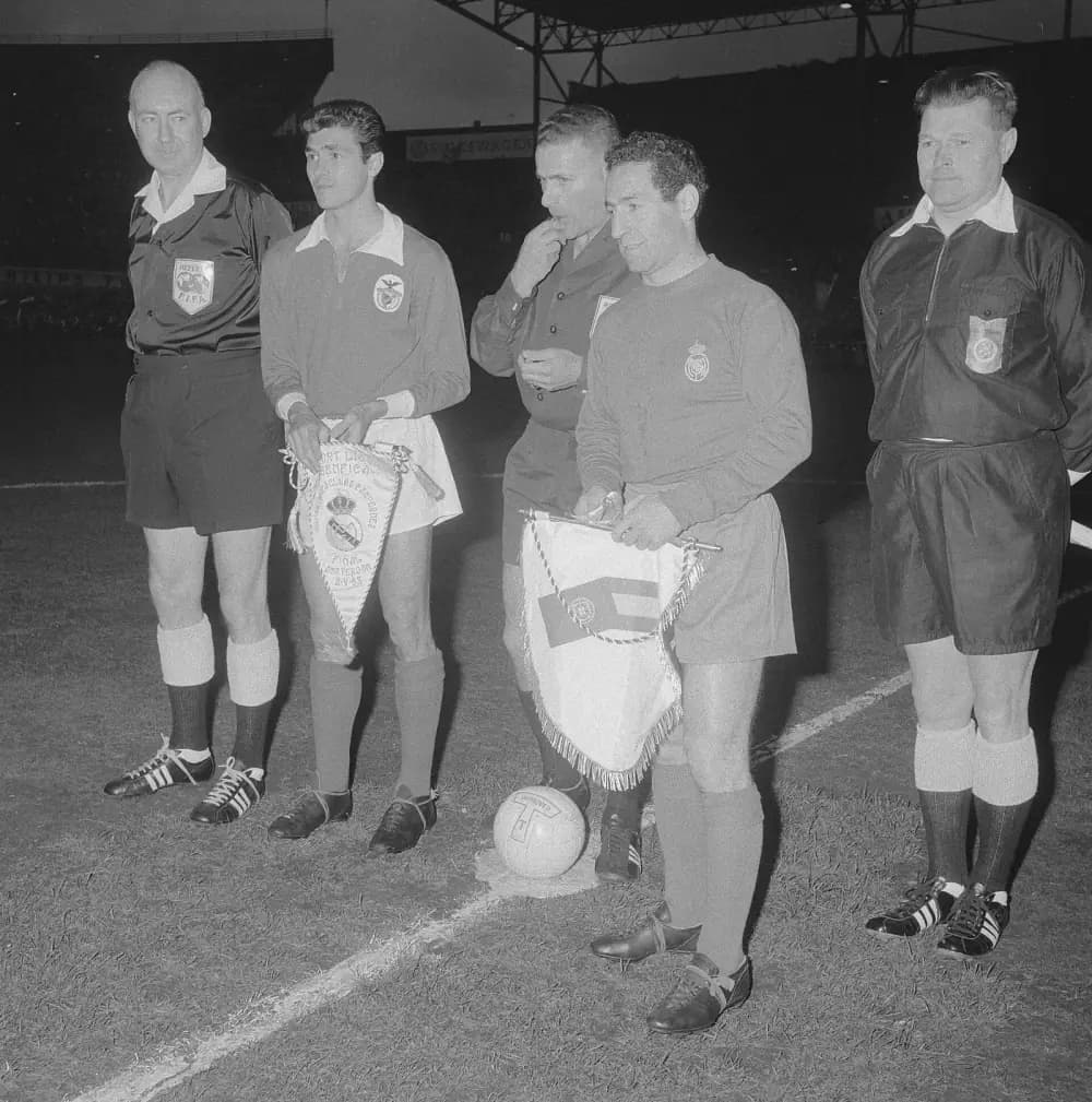 Two footballers, one in a Benfica shirt and one in a Real Madrid shirt, stand on a floodlit pitch holding pennants, flanked by three match officials in black. A ball sits on the centre spot. The pennant held by the Benfica player carries a crowned crest and the words Final Amsterdam 2-V-62.