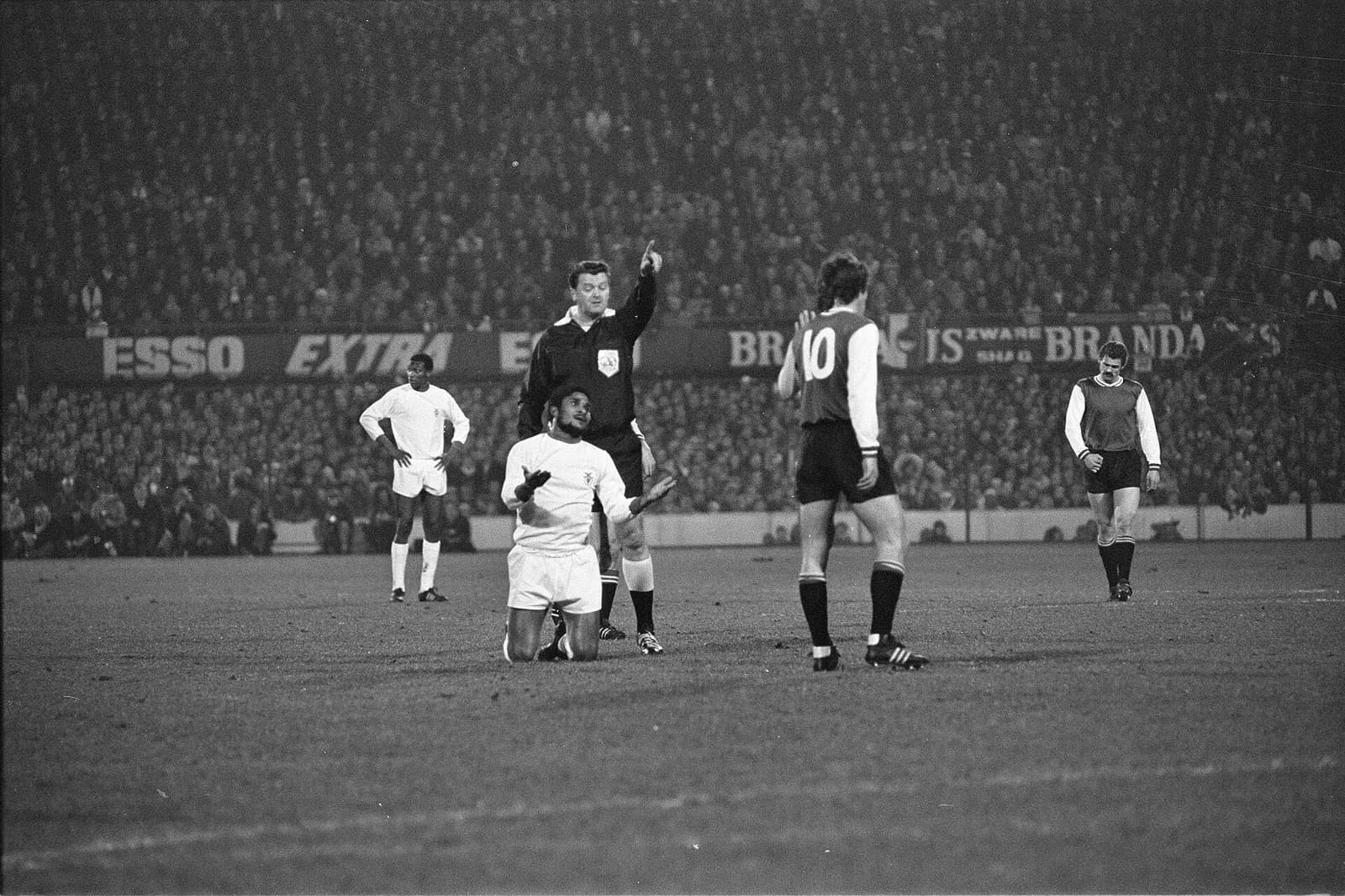 Night match in a packed stadium. A bearded Benfica player in a white kit kneels on the pitch with his palms raised, in front of a referee in black who points into the distance. A Feyenoord player in a two-tone shirt with the number 10 on his back stands beside them.