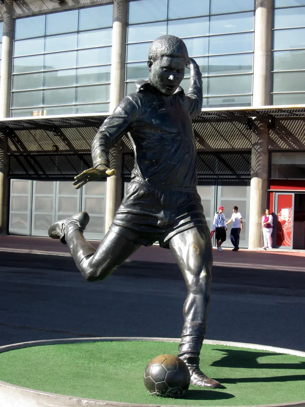 A bronze statue of a footballer in shirt and shorts, right leg drawn back and left hand raised to his head, about to strike a ball. It stands on a round patch of artificial grass in front of the glass and concrete facade of a stadium.