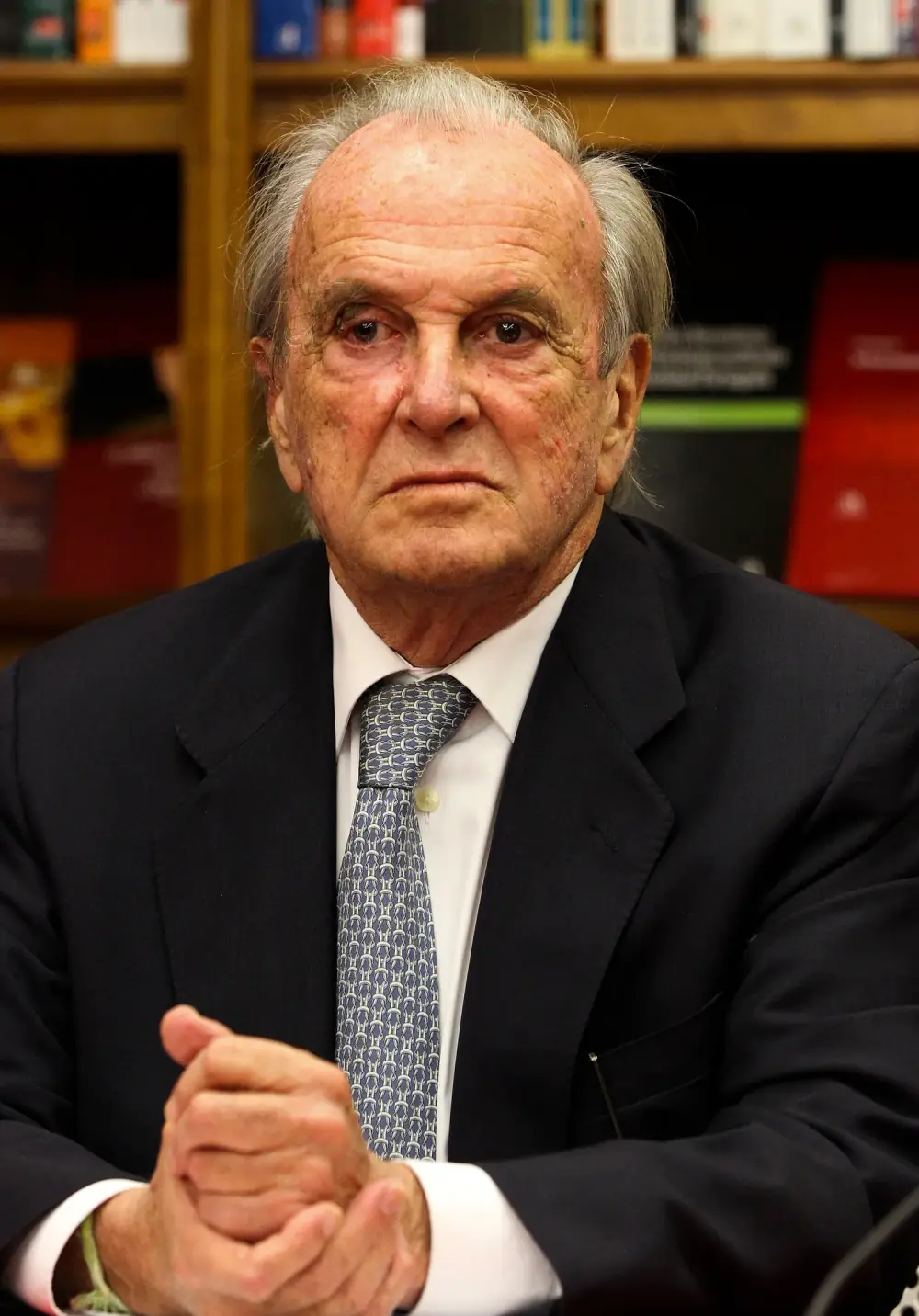An elderly man with swept-back grey hair, in a dark suit, white shirt and patterned blue-grey tie, sits with his hands clasped in front of shelves of books.