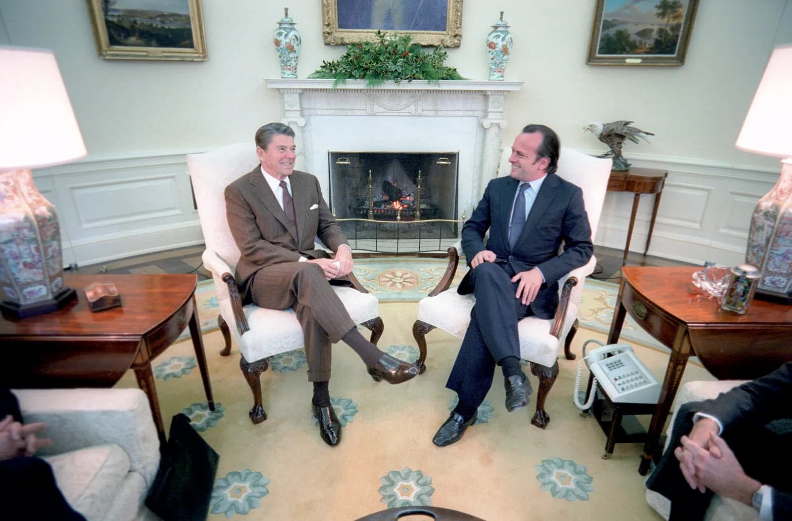 Two men in suits sit in white armchairs on either side of a lit fireplace in the Oval Office, smiling at each other, with holly on the mantelpiece. Ronald Reagan, on the left, wears a brown suit; Francisco Pinto Balsemão, younger and dark-haired, on the right, wears a navy suit and sits with his legs crossed.