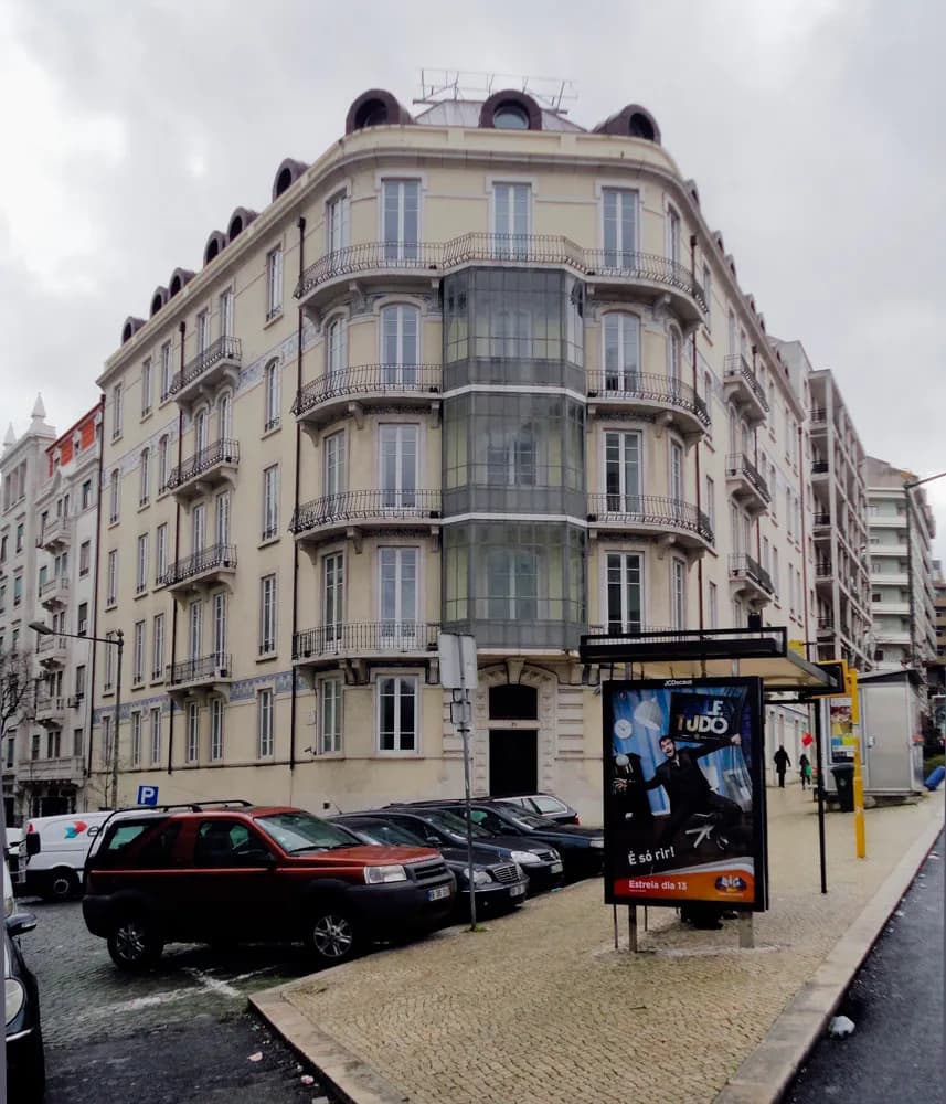 A pale six-storey apartment building on a street corner, with iron balconies, a glazed bay running up the rounded corner and round dormer windows on the roof. Cars are parked in front and a bus shelter stands on the pavement.