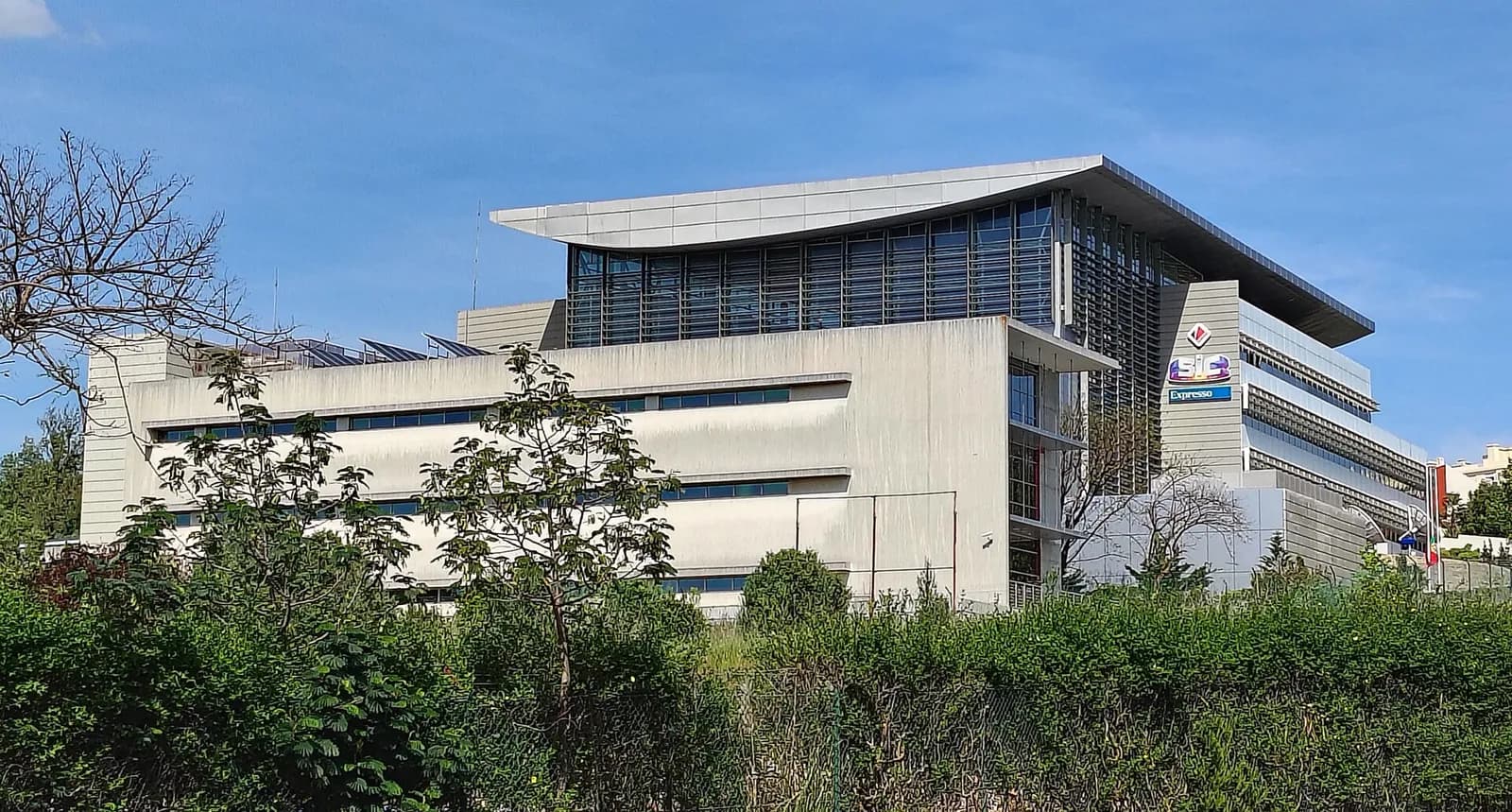 A large modern grey office building with a curved, overhanging roof behind a screen of trees and shrubs, with the SIC logo and an Expresso sign on a tower at one corner.