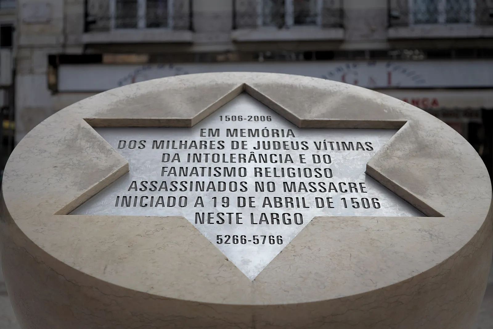 Close-up of a pale stone drum with a six-pointed star cut into its top, bearing an engraved Portuguese inscription and the dates 1506–2006 and 5266–5766, in front of a city building.