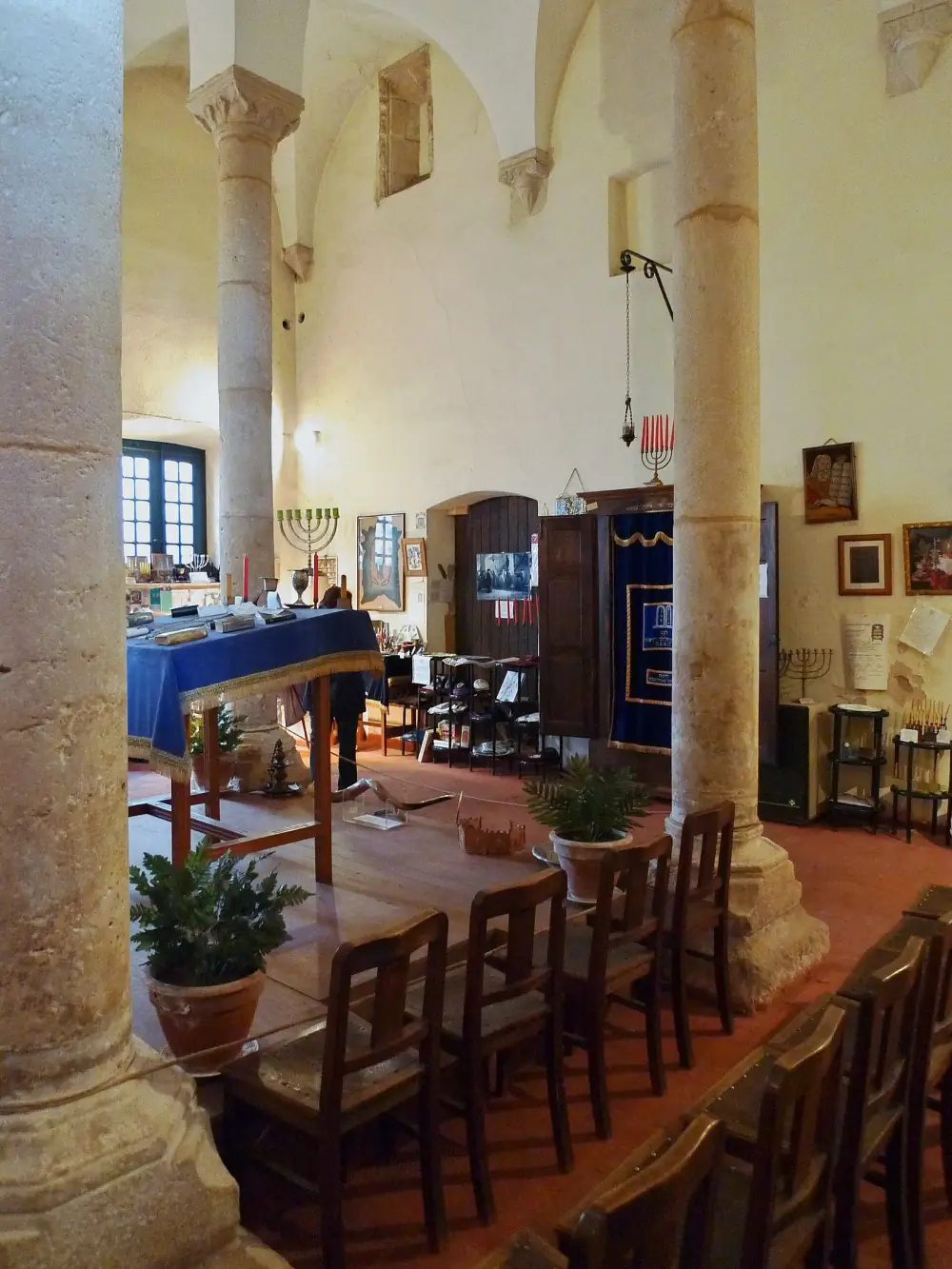 Inside a whitewashed hall with a vaulted ceiling held up by stone columns, wooden chairs face a reading table covered in a blue cloth, with branched candlesticks and an ark behind a blue embroidered curtain.