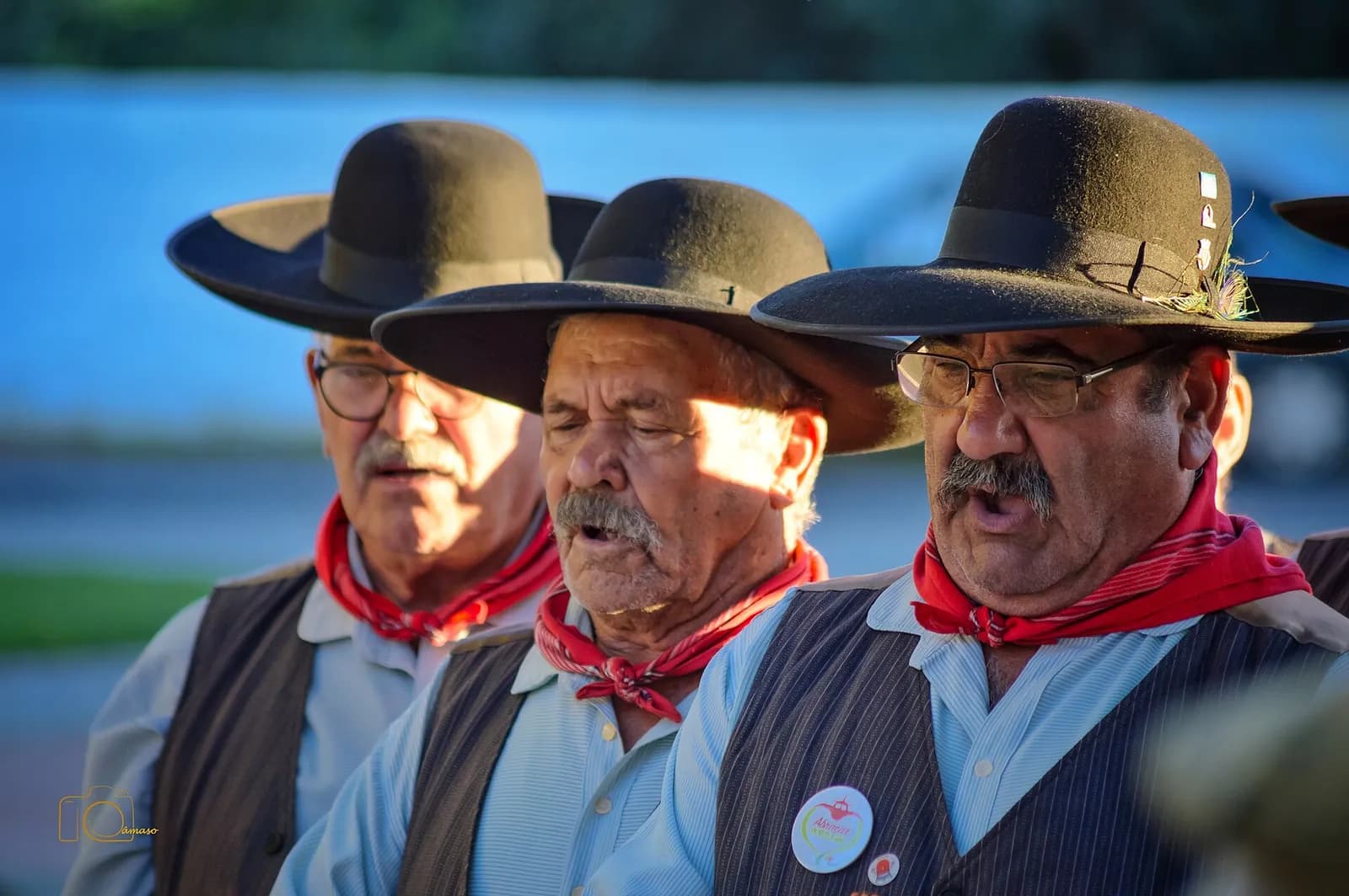 Close-up of three older men singing in a row, wearing black felt hats with wide brims, red neckerchiefs, light shirts and dark waistcoats. The man in the middle sings with his eyes closed; sunlight falls across his face.