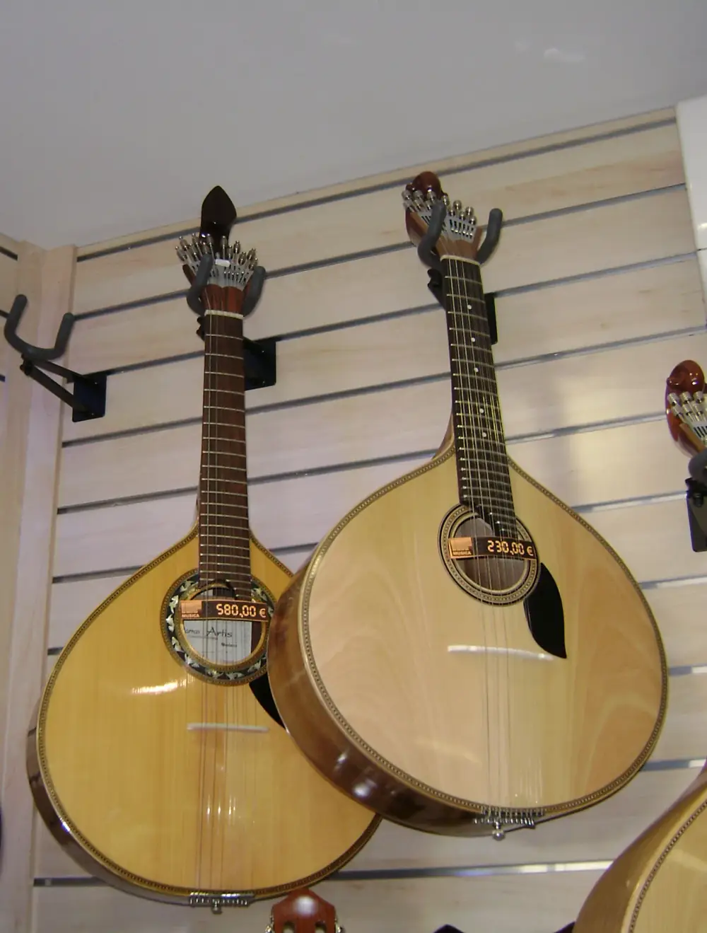 Two pear-shaped twelve-string guitars hang side by side on a shop wall, each with a fan of metal tuning screws at the head. The one on the left has a pointed, teardrop-shaped head; the one on the right has a scroll-shaped head and a slightly rounder body.