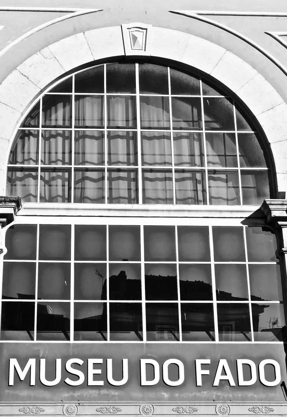 Black and white photograph of a building front: a large semicircular window divided into small panes, set in a stone arch, above a row of square panes and a band with the words MUSEU DO FADO in large capitals.