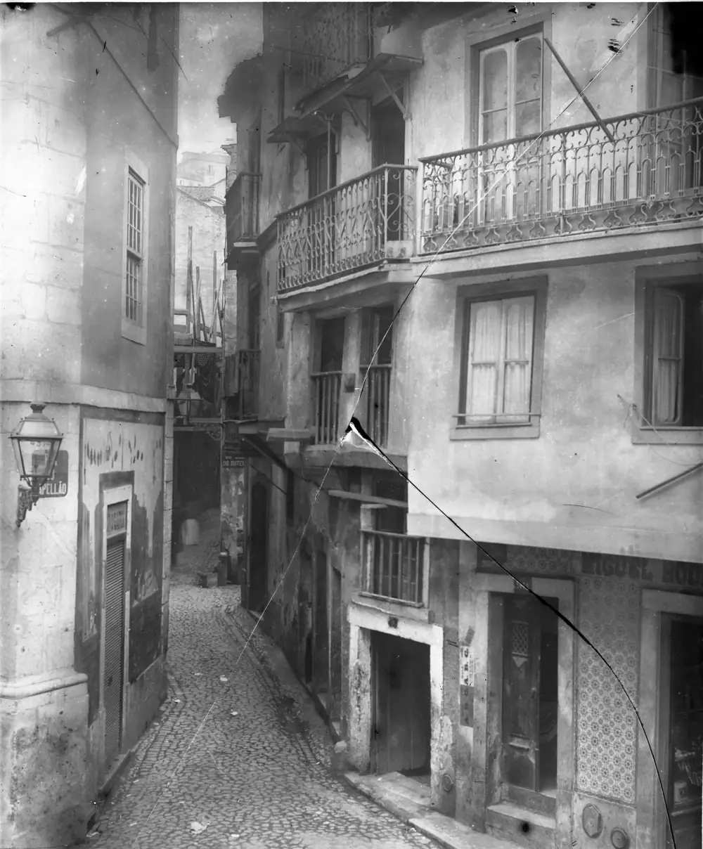 Black and white photograph of a narrow cobbled lane climbing between tall, plain houses with iron balconies. A street lamp and a name plate ending in “apellão” are fixed to the corner building on the left; tiled shopfronts line the right. The glass negative is cracked diagonally.