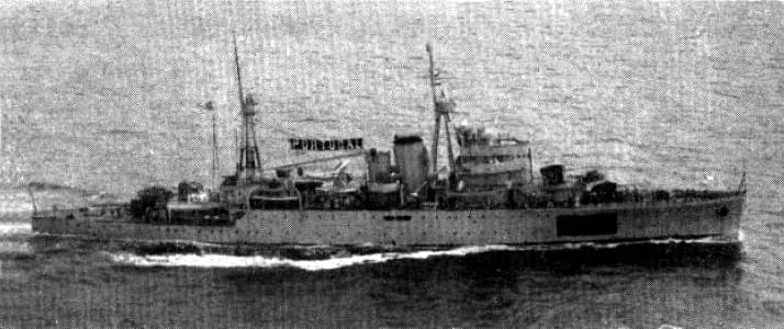 Black-and-white photograph of a long grey warship with two masts and a single funnel cutting through the sea at speed, a bow wave curling along her side.