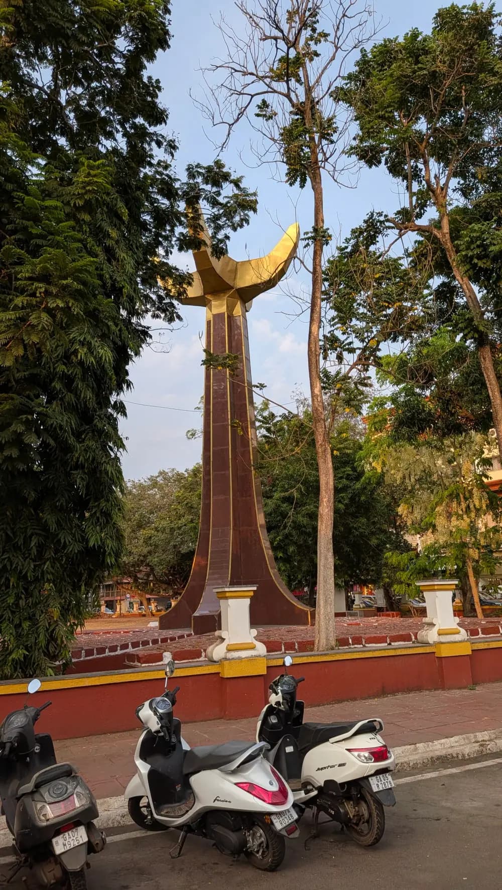 A tall maroon column edged in gold, topped by a gilded sculpture shaped like an opening flower or flame, standing in a small fenced park among trees, with scooters parked on the street in front.