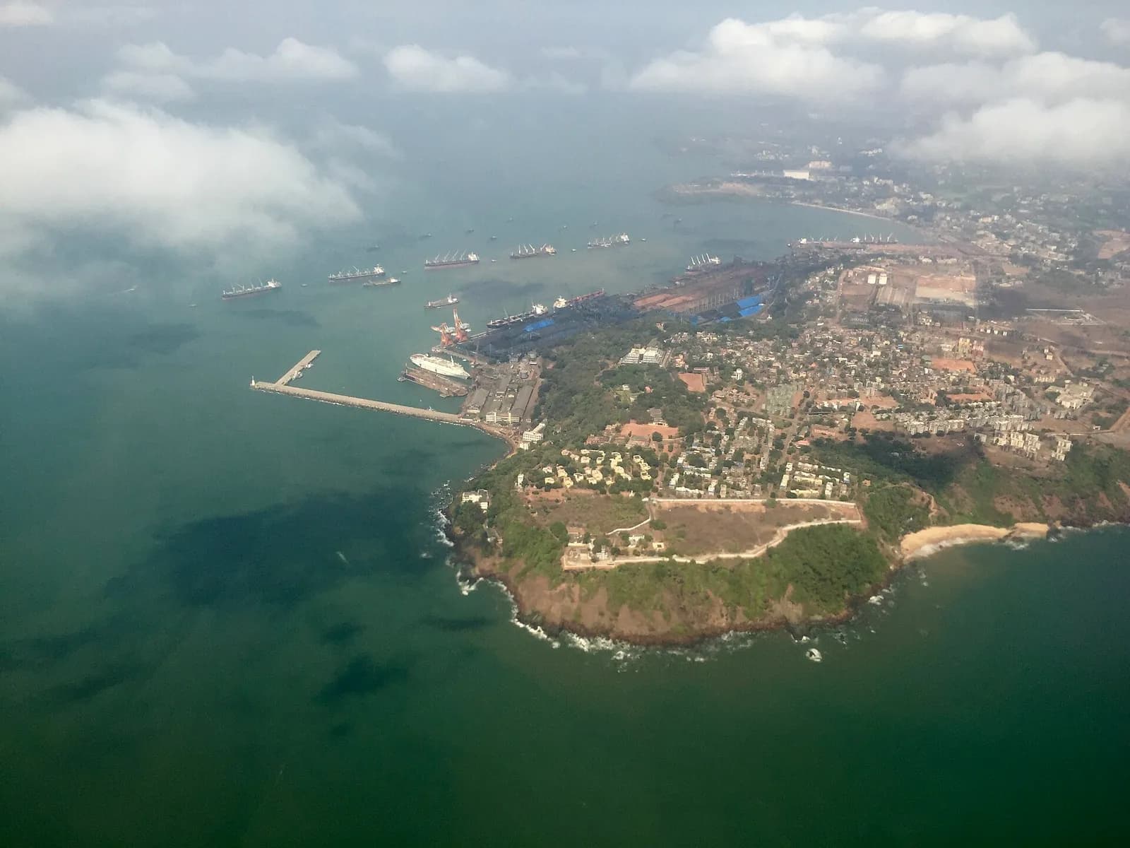 Aerial view of a green headland jutting into the sea, with a long breakwater, a port crowded with cranes and docks, and a line of cargo ships anchored in the bay beyond.