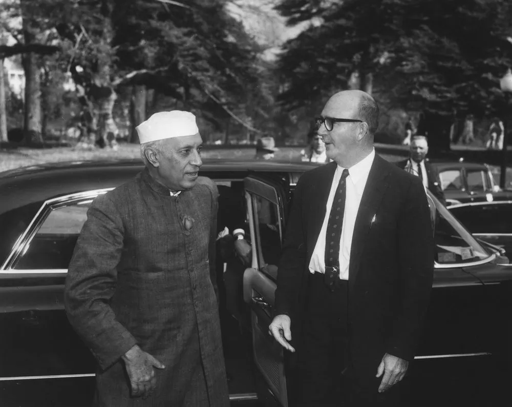 Black-and-white photograph of Jawaharlal Nehru in a white cap and long buttoned coat beside the open door of a large black car, greeted by a tall bald man in glasses and a dark suit, with trees behind them.