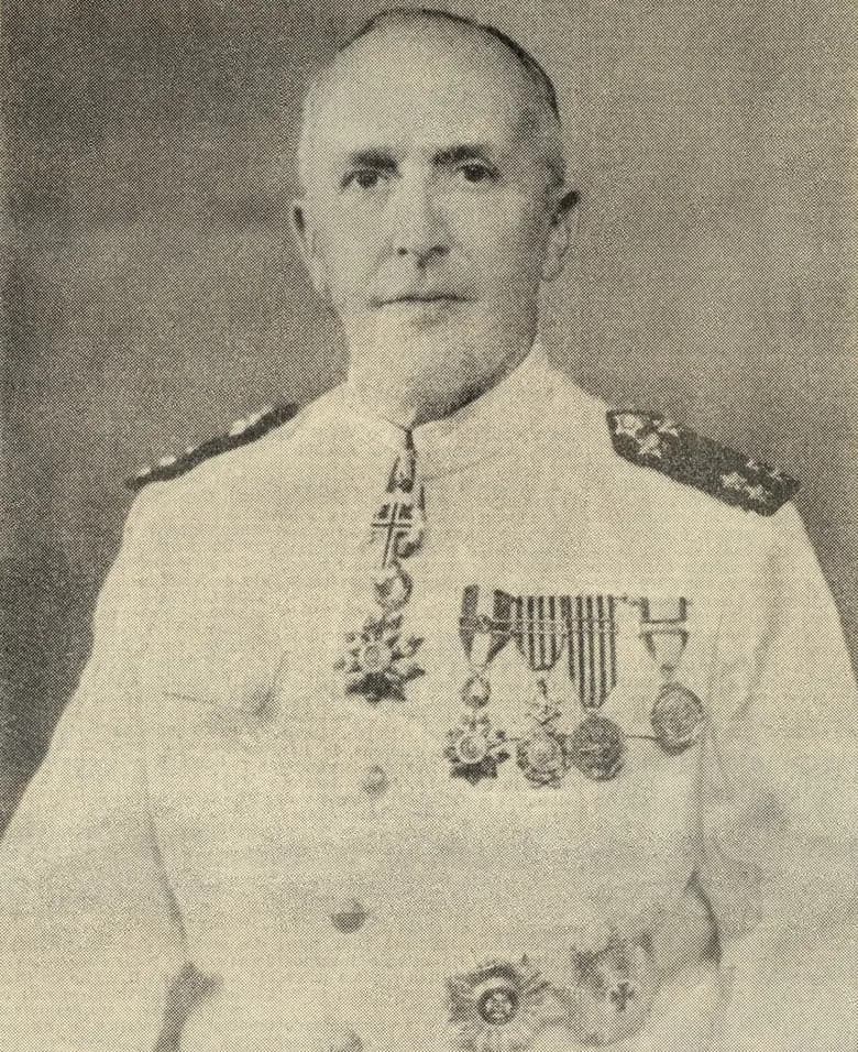 Half-length black-and-white portrait of a grey-haired, clean-shaven man in a white military tunic with dark shoulder boards, a cross on a ribbon at his collar and a row of medals on his chest.