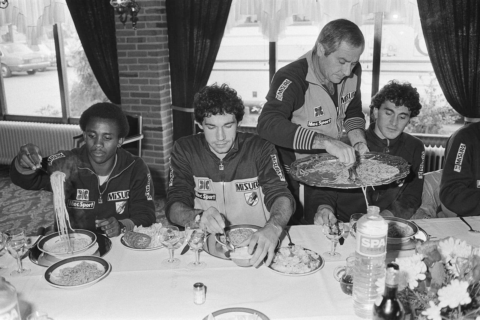 Black-and-white photo of footballers in tracksuits eating spaghetti at a long table set with glasses and a bottle of water. A grey-haired man stands behind them serving pasta from a silver platter. On the left a young Black player lifts a forkful of spaghetti.