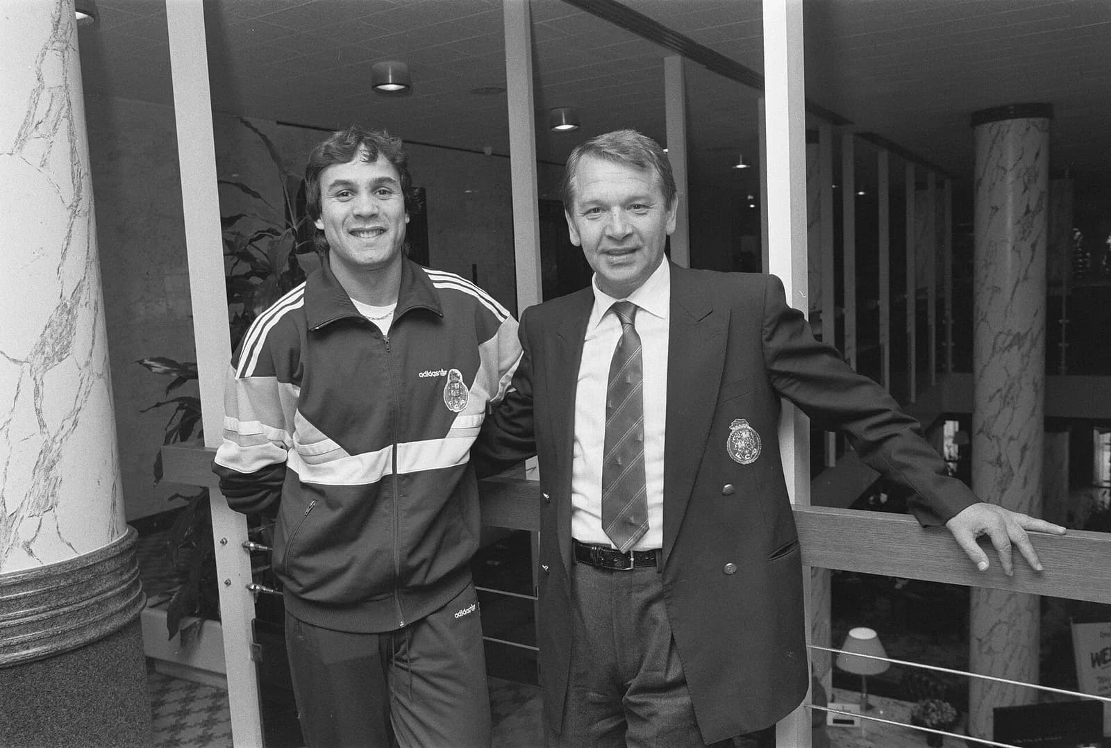 Black-and-white photo of two men standing by a railing in a hotel lobby. On the left a smiling young man with dark wavy hair wears an Adidas tracksuit top with the FC Porto crest; on the right an older man in a club blazer with the same crest, shirt and striped tie leans on the rail.