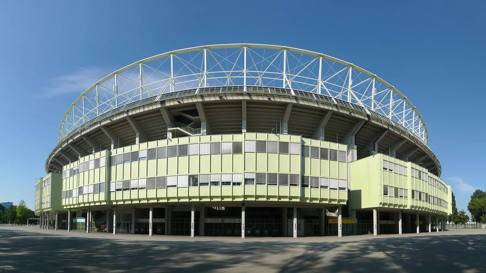 Wide-angle view of a large oval stadium under a clear blue sky. Pale green office blocks wrap the base, the curved concrete stands rise behind, and a white steel truss roof frame arches over the top.