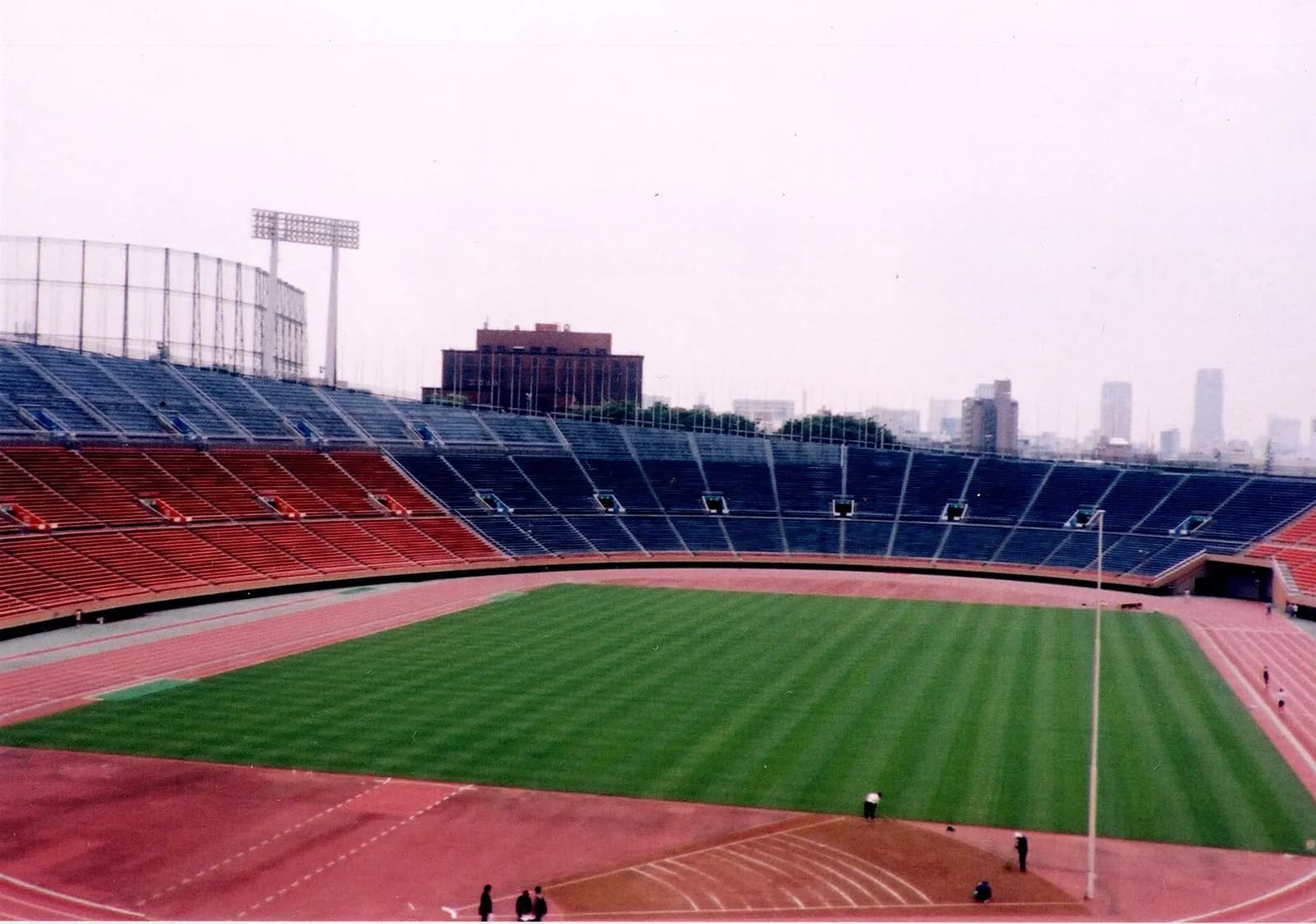 An athletics stadium, empty but for a few groundstaff, seen from high in the stands on an overcast day: a striped green football pitch inside a red running track, with tiers of red and blue seats beyond and city buildings on the horizon.