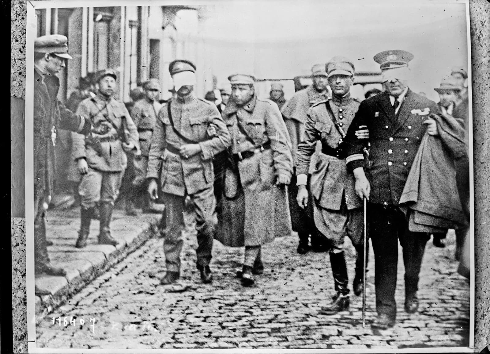 Two blindfolded officers are led arm in arm along a cobbled street by soldiers. One wears army uniform; the other, on the right, wears a dark naval uniform and carries a cane and a folded cloak. Troops and onlookers follow behind.