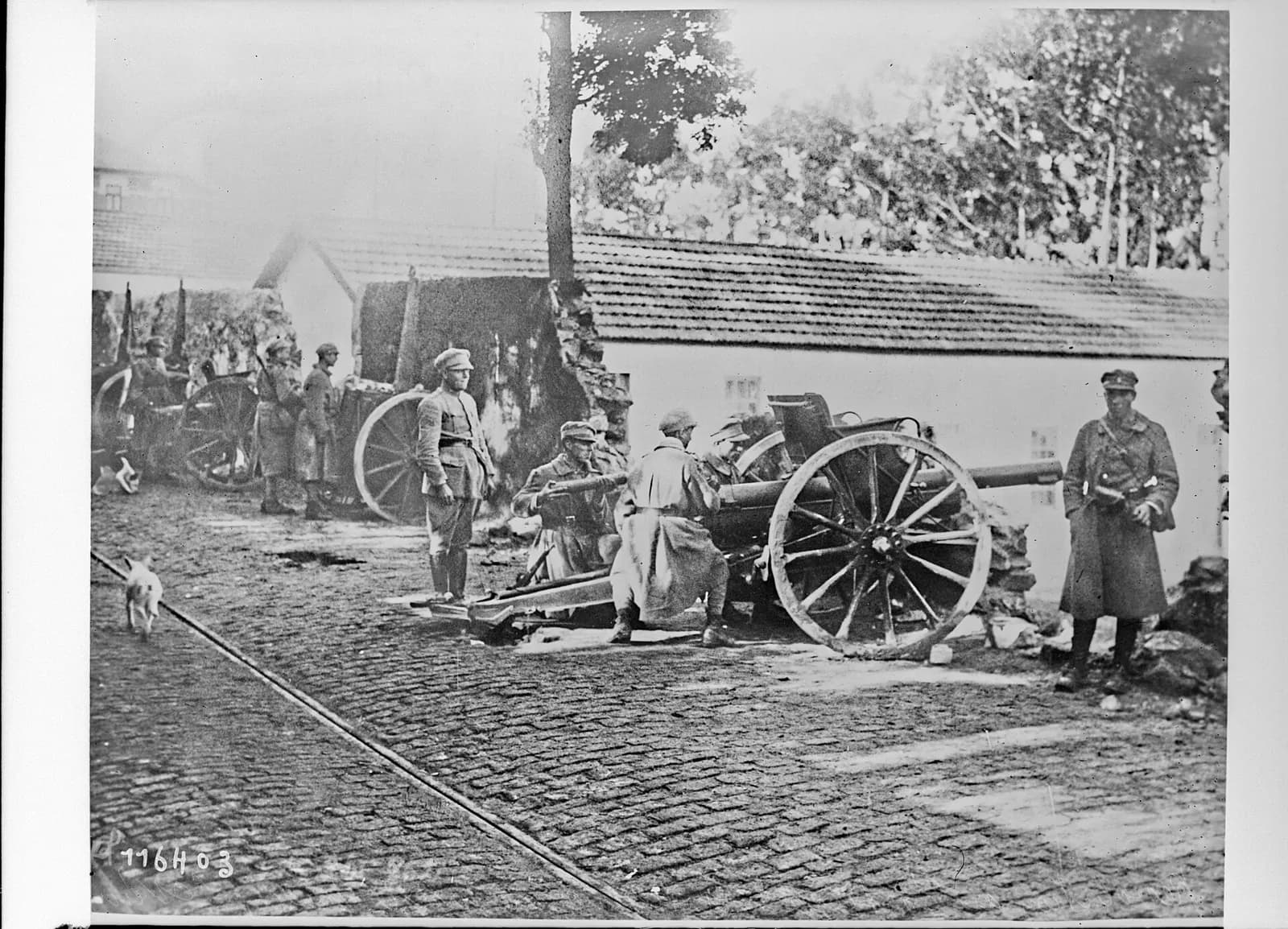 Soldiers kneel to load a field gun with large spoked wheels on a cobbled road beside a low tiled-roof building. More guns, soldiers and a small dog are behind them.