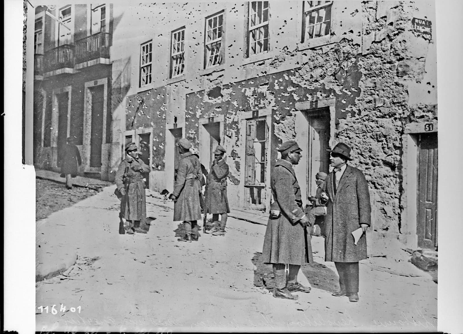 Soldiers in greatcoats and a man in a hat and overcoat talk in a sloping street in front of a house whose walls are pitted with bullet holes and patches of fallen plaster, its windows broken.