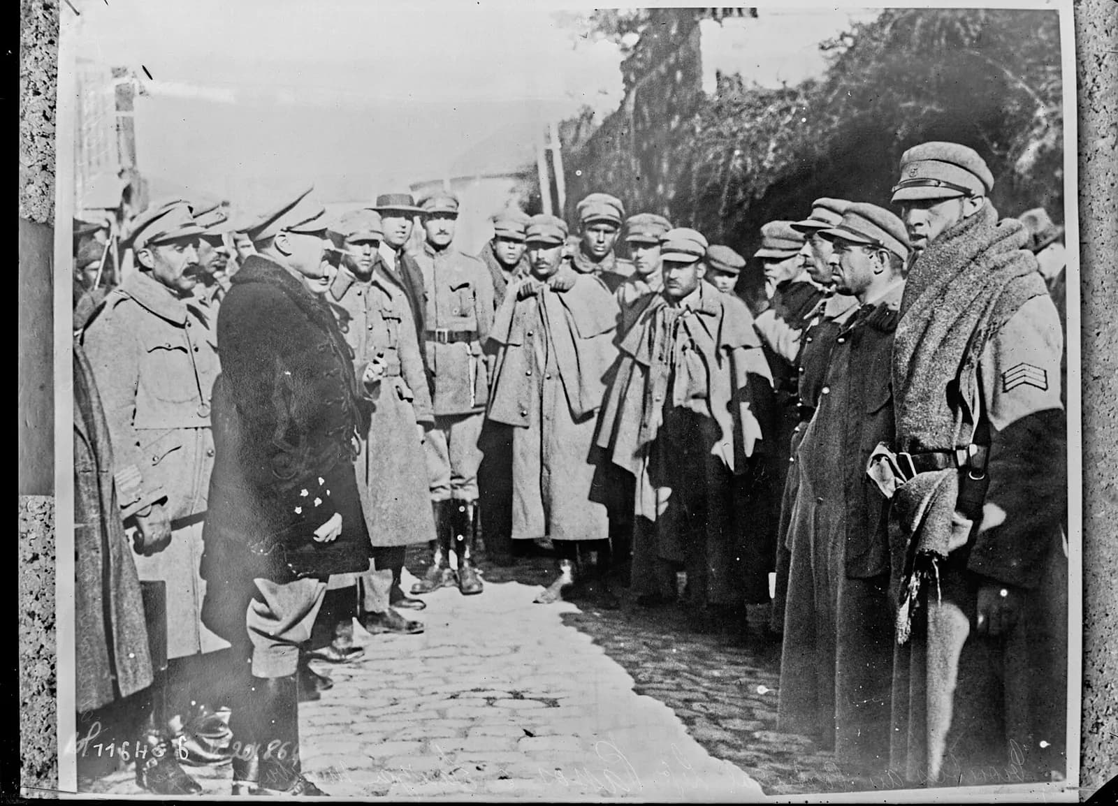 An officer in a dark, fur-trimmed coat and peaked cap stands on a cobbled path talking to a line of soldiers in greatcoats and capes, some with blankets around their shoulders.