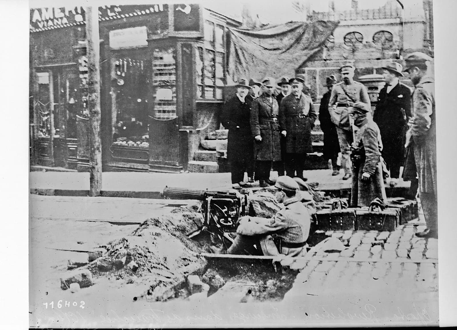 A soldier crouches behind a heavy machine gun in a trench dug into a cobbled street. Officers and men in overcoats and hats stand on the pavement behind him, in front of shop windows.