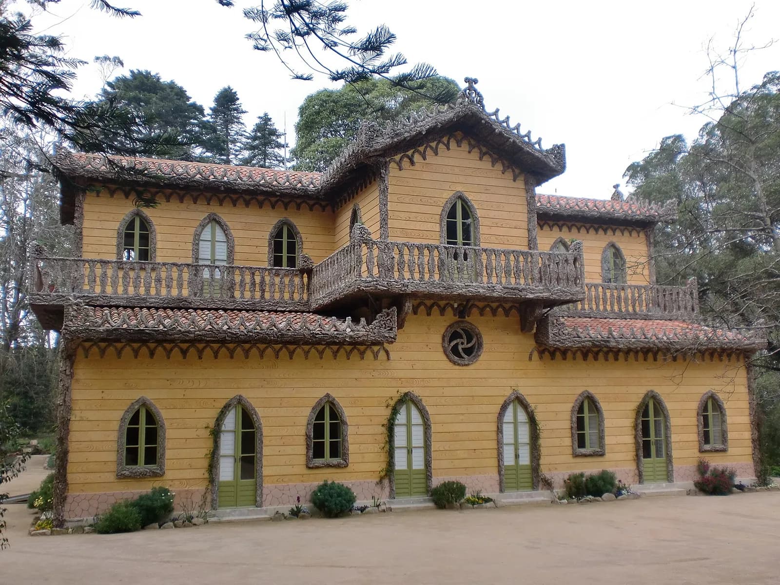 A two-storey ochre house with dark brown trim, pointed arched doors and windows, wooden balconies and deep eaves, set on a gravel yard among tall trees.