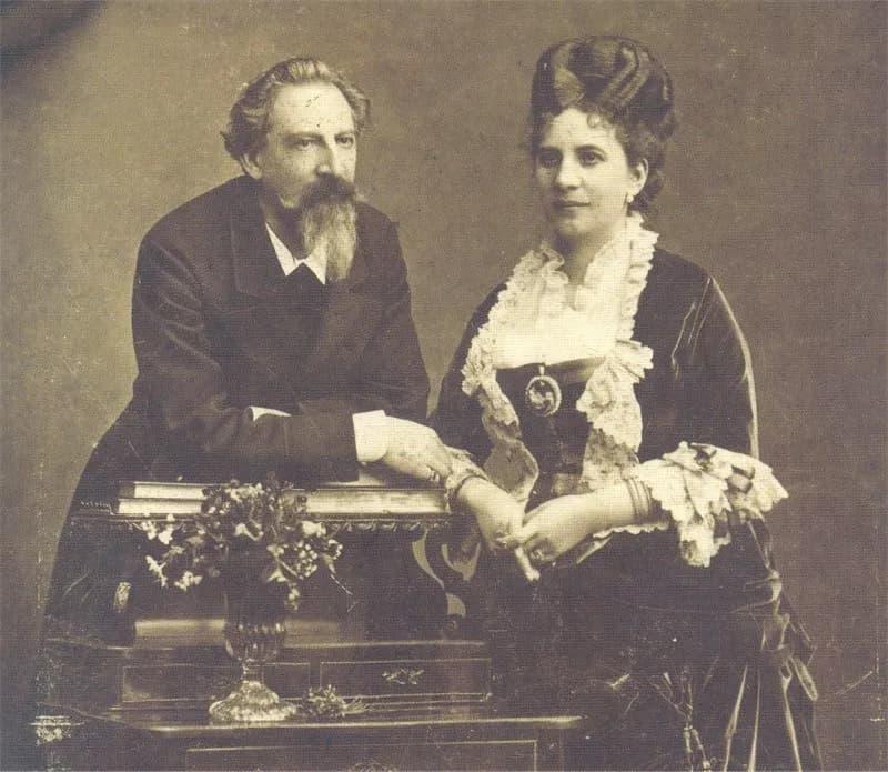 Sepia photograph of a bearded older man in a dark suit leaning on a small table beside a dark-haired woman in a dark dress with a wide lace collar; their hands meet on the table next to a vase of flowers.