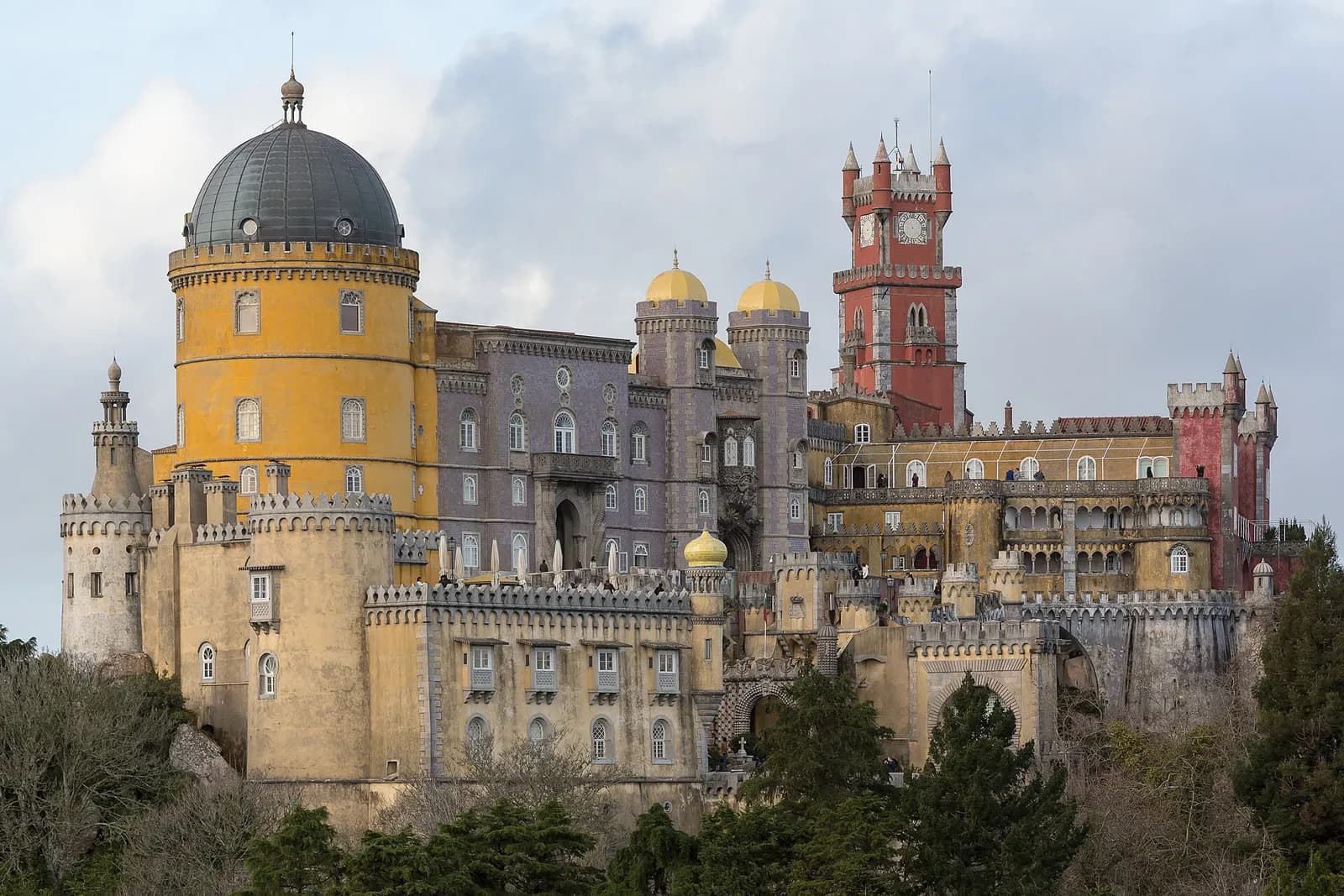 A hilltop palace rises above dark woodland under a cloudy sky: a round ochre-yellow tower with a grey dome on the left, a lilac-tiled block with two small yellow domes in the middle and a tall red clock tower on the right, all above crenellated ochre walls.