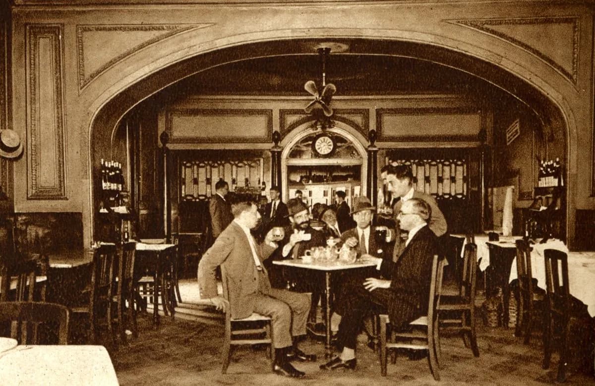 A sepia photograph of a café interior under a wide arch. Five men in suits, two of them in hats, sit and stand around a small marble table with a teapot and cups; a clock hangs over a doorway behind them.