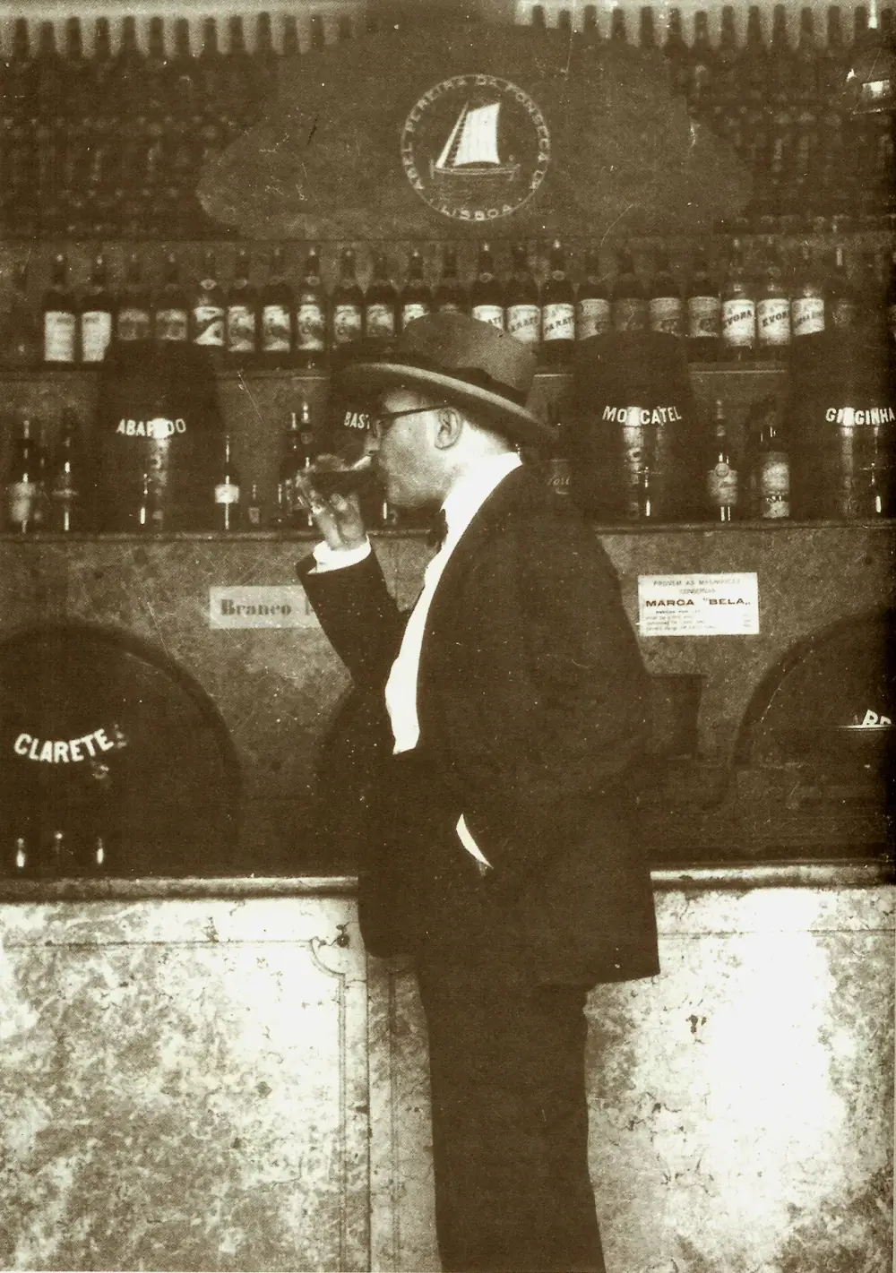 A man in a hat, round glasses, bow tie and dark suit stands in profile at a shop counter, raising a small glass to his lips. Behind him are shelves of bottles and barrels labelled Abafado, Moscatel, Ginginha and Clarete under a sign reading Abel Pereira da Fonseca, Lisboa.