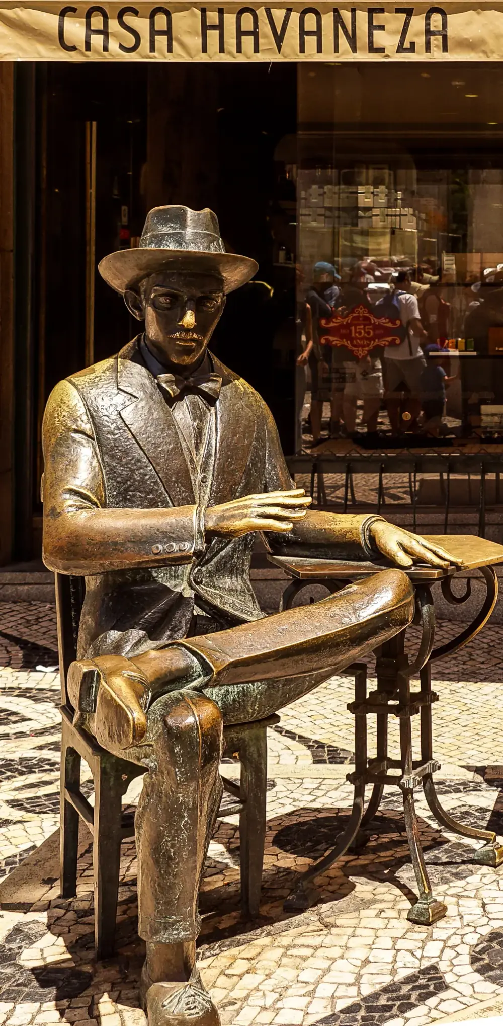 A bronze statue of a thin man in a hat, bow tie and three-piece suit sits cross-legged on a bronze chair beside a small café table, on a black-and-white cobbled pavement in front of a shop sign reading Casa Havaneza.
