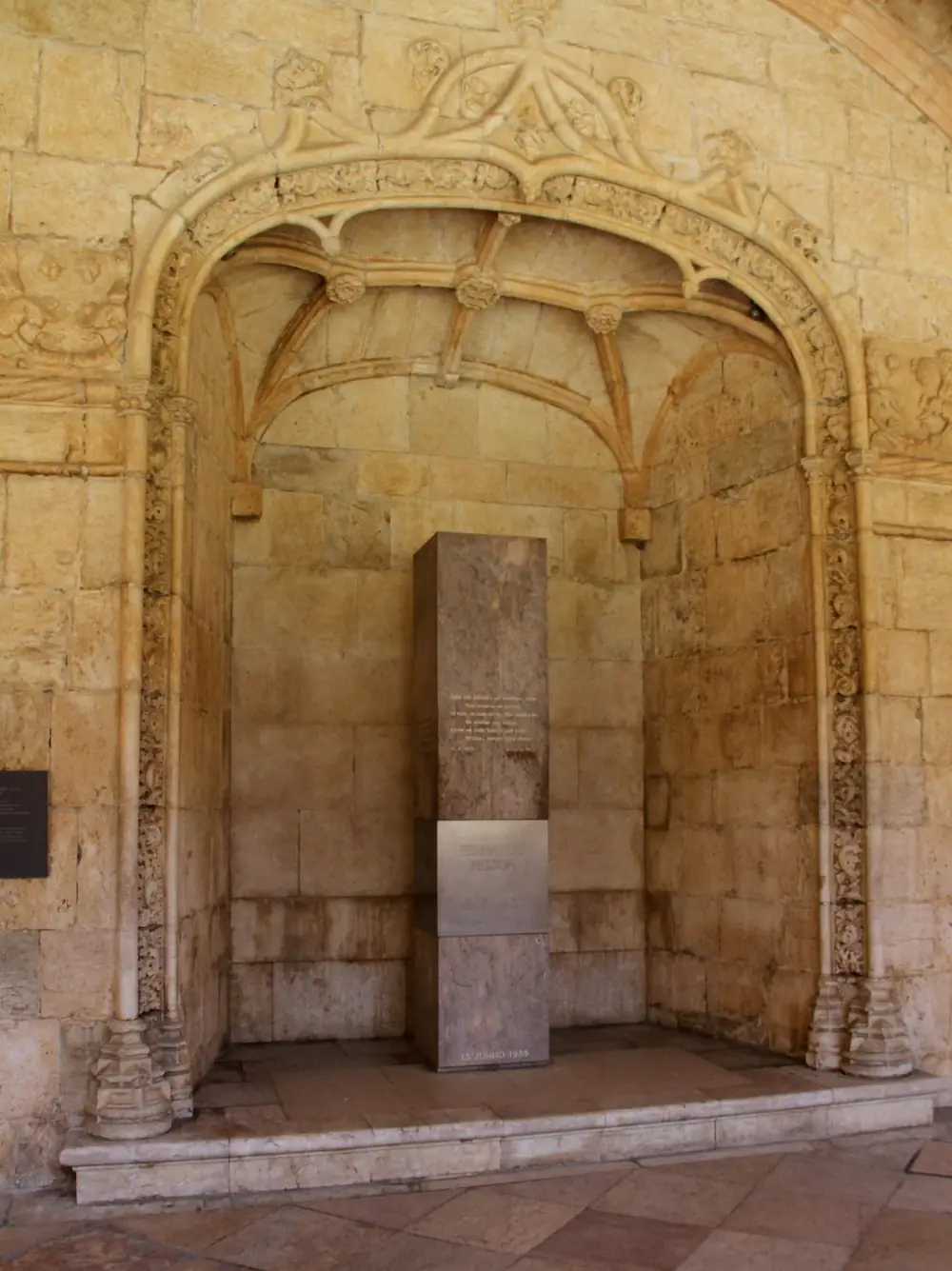 A tall square pillar of polished stone with a band of brushed steel stands alone in a vaulted niche of carved honey-coloured limestone. Lines of verse are engraved on the upper stone and Fernando Pessoa's name on the steel.