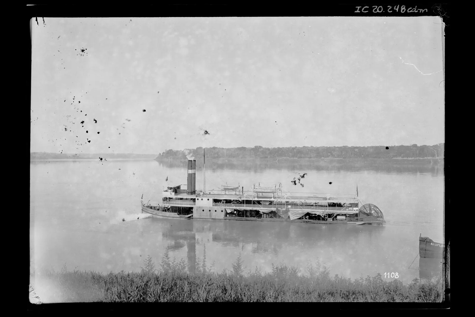 Black-and-white photograph of a two-deck river steamer with twin funnels and a paddle wheel at the stern, crossing a wide, still river. A low line of forest runs along the far bank and tall grass fills the foreground.