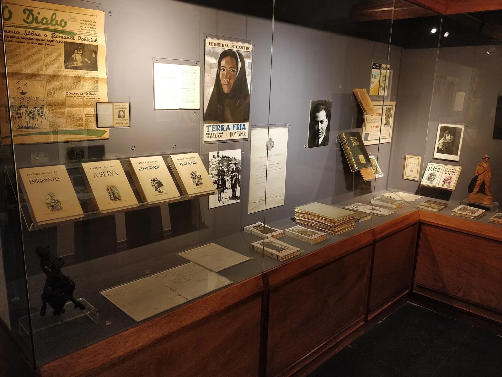 A glass museum case holding four matching volumes titled Emigrantes, A Selva, Eternidade and Terra Fria, with a yellowed front page of the newspaper O Diabo, a poster for Terra Fria showing a woman in a dark hood, photographs and documents on the wall behind.