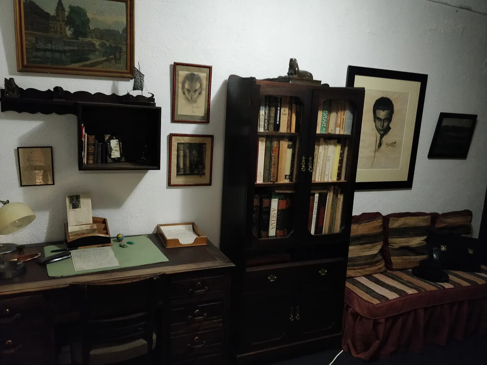 A small room with a wooden writing desk and chair, a glass-fronted bookcase full of books, a striped daybed, and framed drawings and paintings on whitewashed walls, among them a charcoal portrait of a young man.