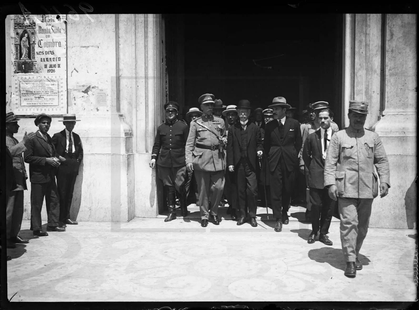A black and white photograph of a group of men leaving the doorway of a stone building. In the centre an officer in uniform walks beside an older man in a dark suit and hat; soldiers and men in boaters stand to the sides.
